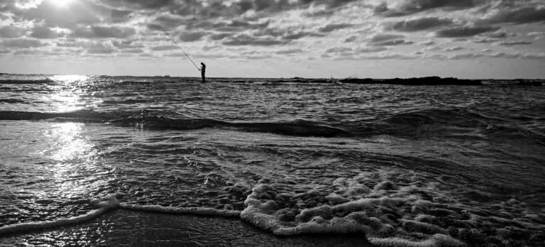Schwarz-weiße Ansicht eines Strandes mit sanften Wellen und einem Leuchtturm im Hintergrund.